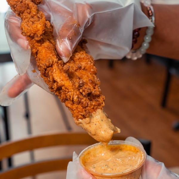 A hand in a clear glove dips a crispy fried chicken tender into a cup of creamy sauce.