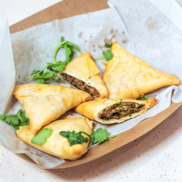Golden-brown, triangular pastries filled with savory meat and peas, garnished with fresh cilantro in a paper tray.