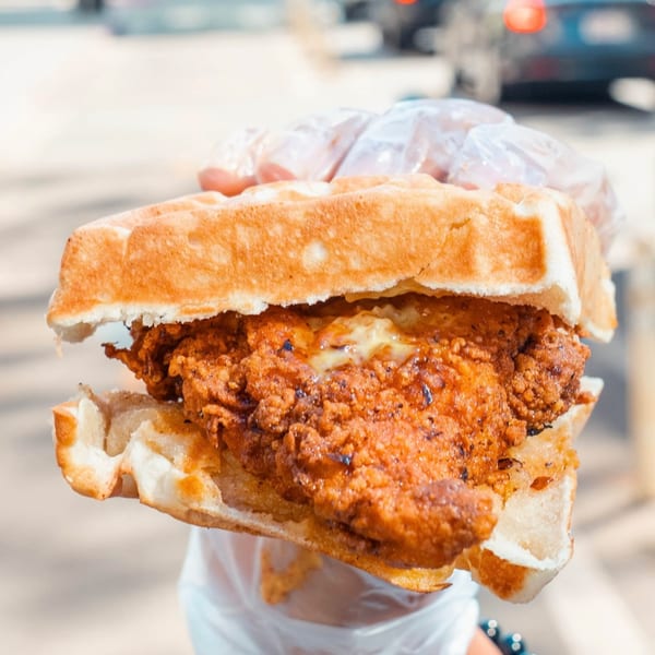 A hand holds a crispy fried chicken sandwich between two golden waffles from Ali's Chicken & Waffles.