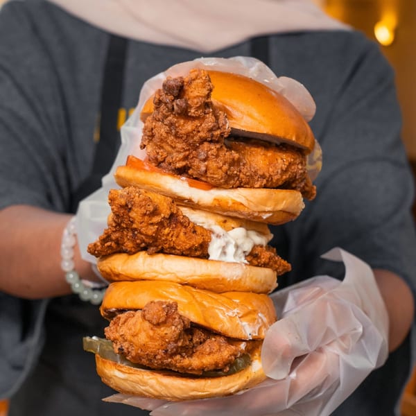 Hands in clear gloves hold a towering stack of three crispy fried chicken sandwiches.